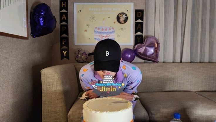 This contains an image of Jin wearing a black face cap and a cake in front of him