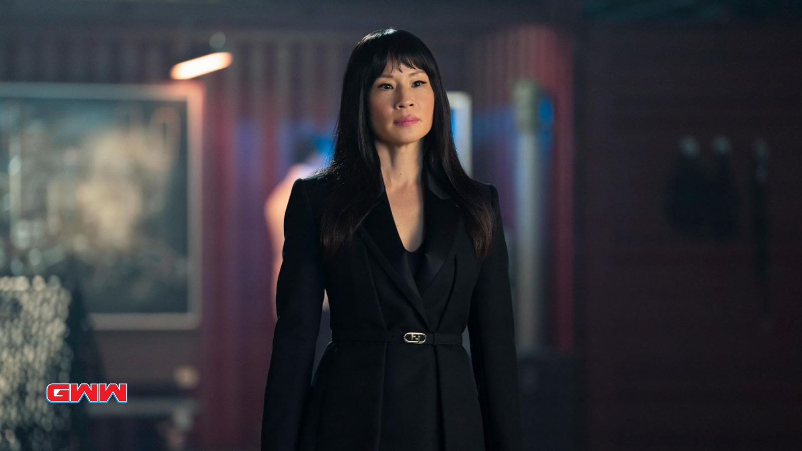 Zoe in a black suit stands confidently in a dimly lit room.