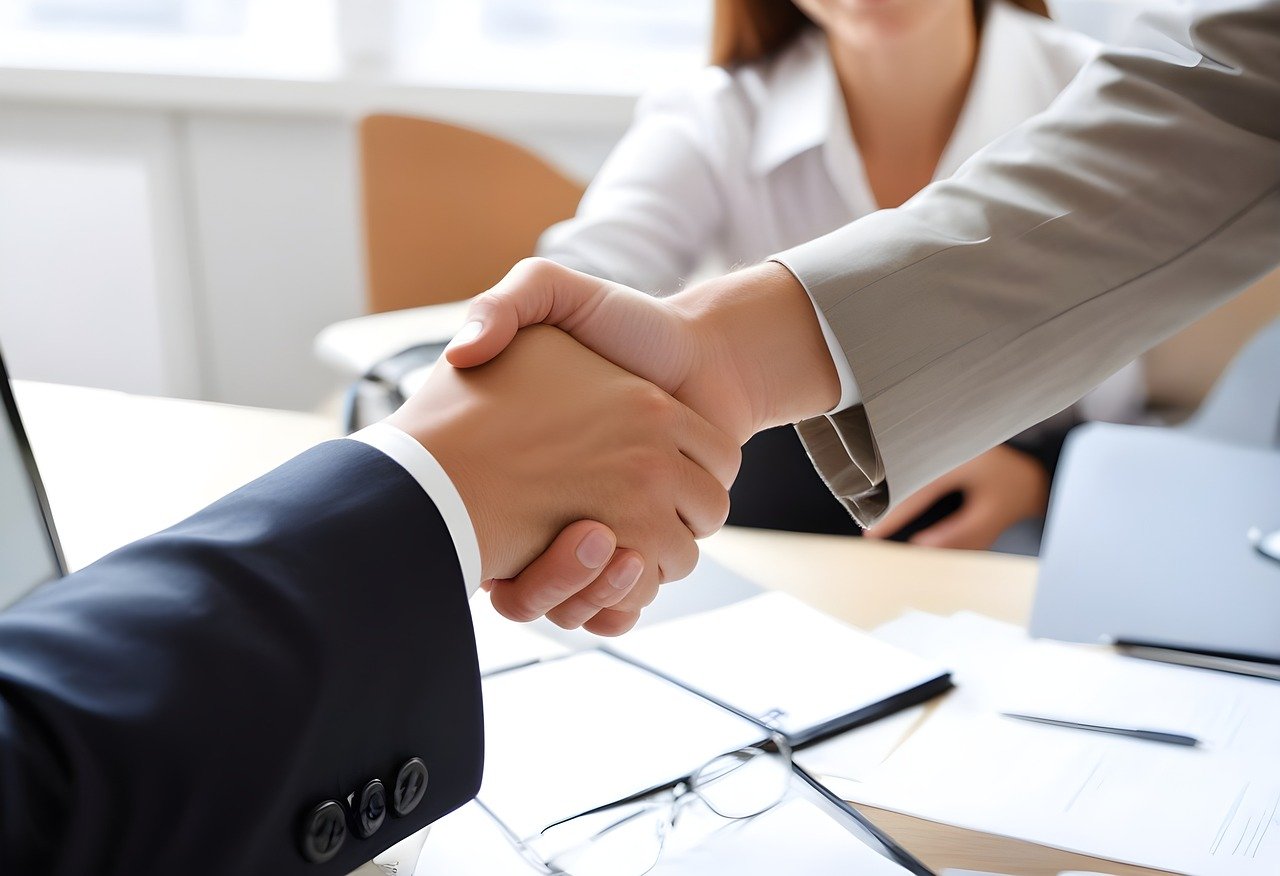 A close-up of two people in business attire shaking hands across a desk with papers and a pair of glasses in view.