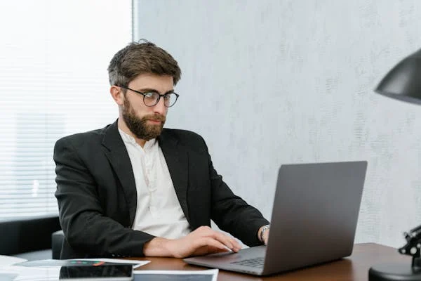 Corporate employee working on laptop wearing black suit and eyeglasses considering factors for rejecting a job proposal