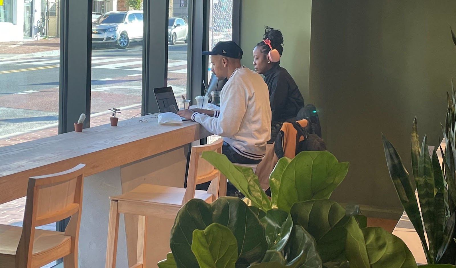 A man on a lap top and a woman with headphones sit at a window table in a cafe with large plants.