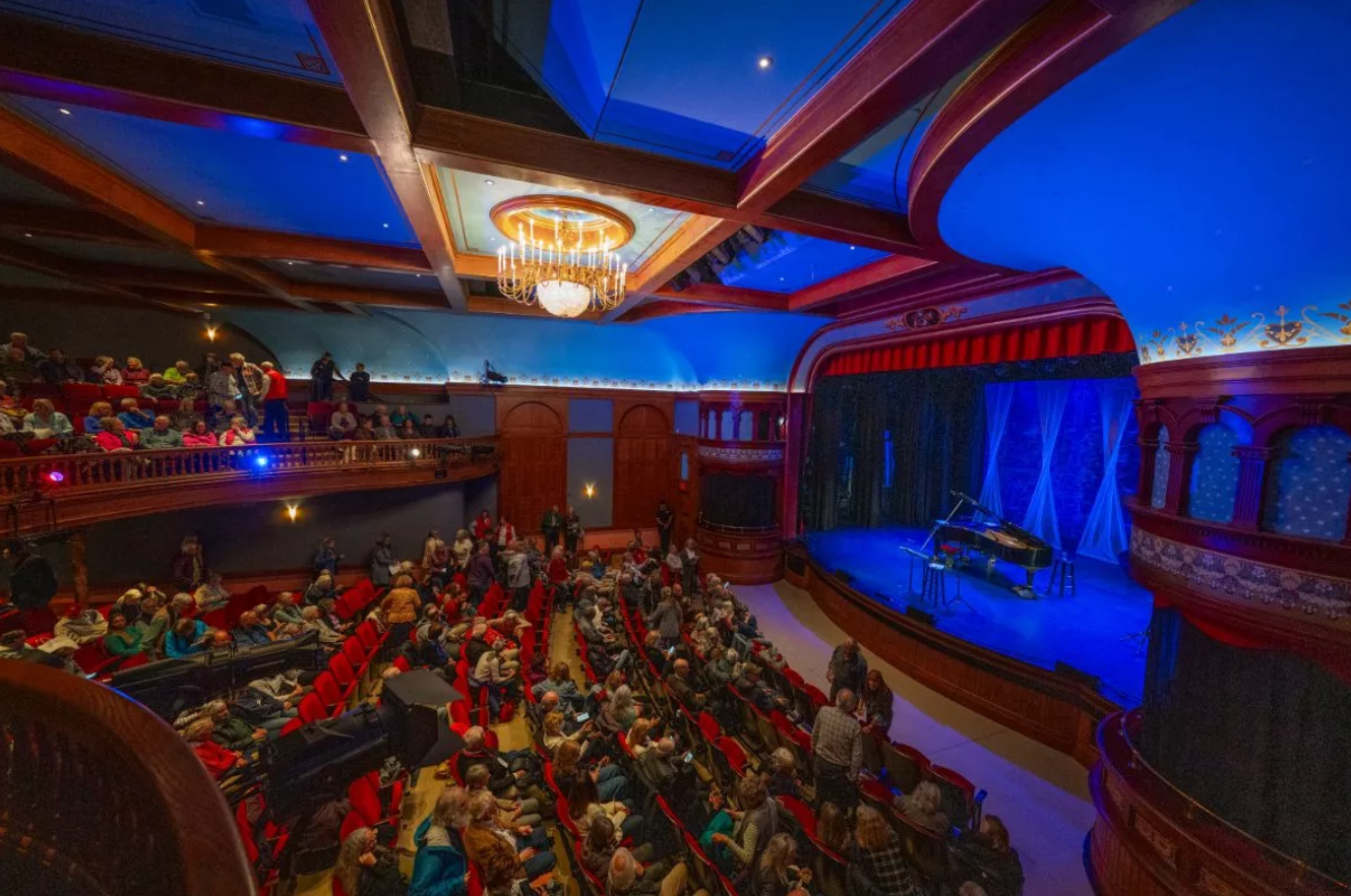Interior of the Wheeler Opera House in Aspen, Colorado, featuring a curved balcony, red velvet seats, chandeliers and a painted proscenium stage set against blue and wood-paneled walls.