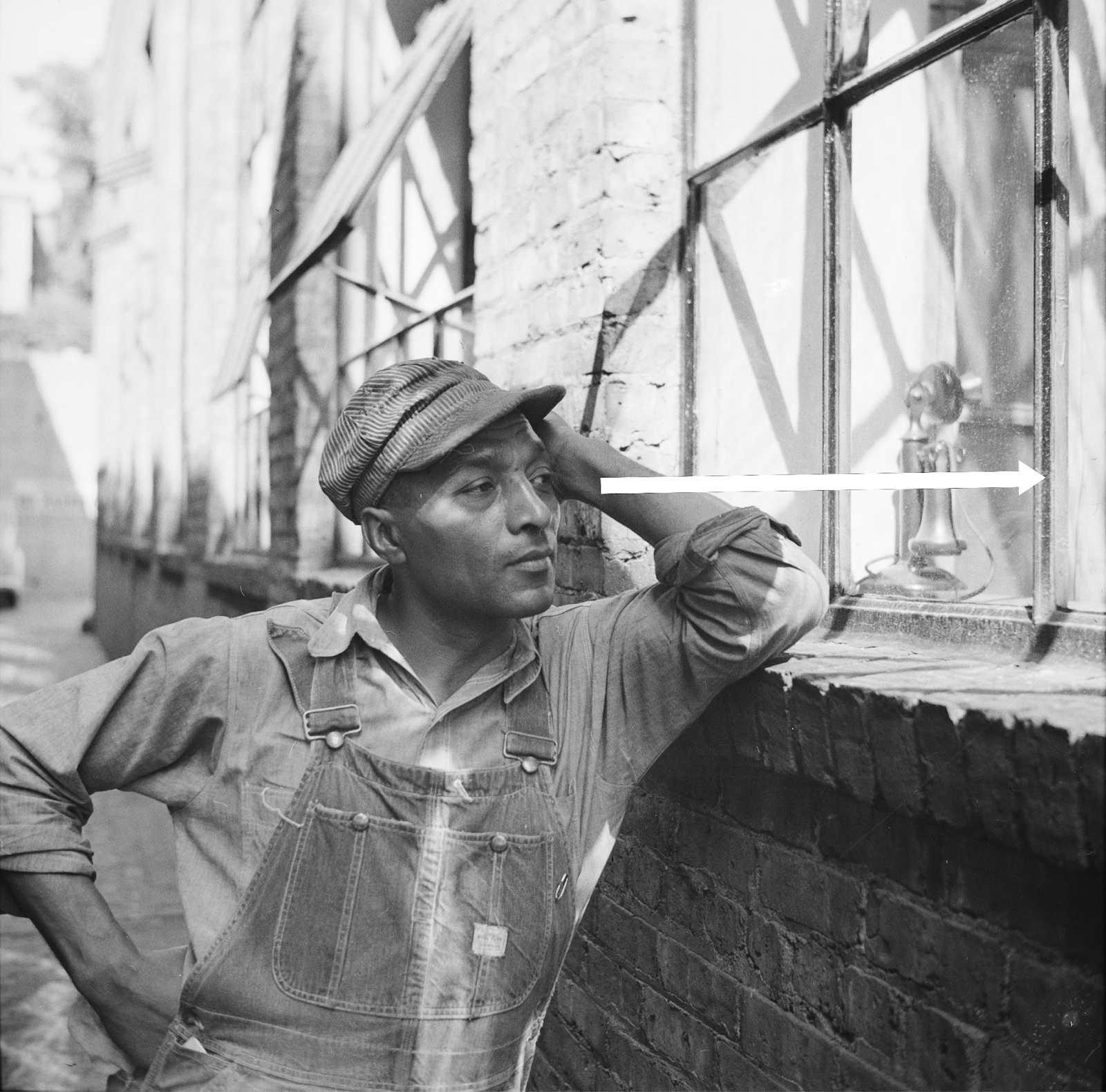 A man in a cap and overalls gazing in a window, leaning his elbow on the brick frame beneath the window