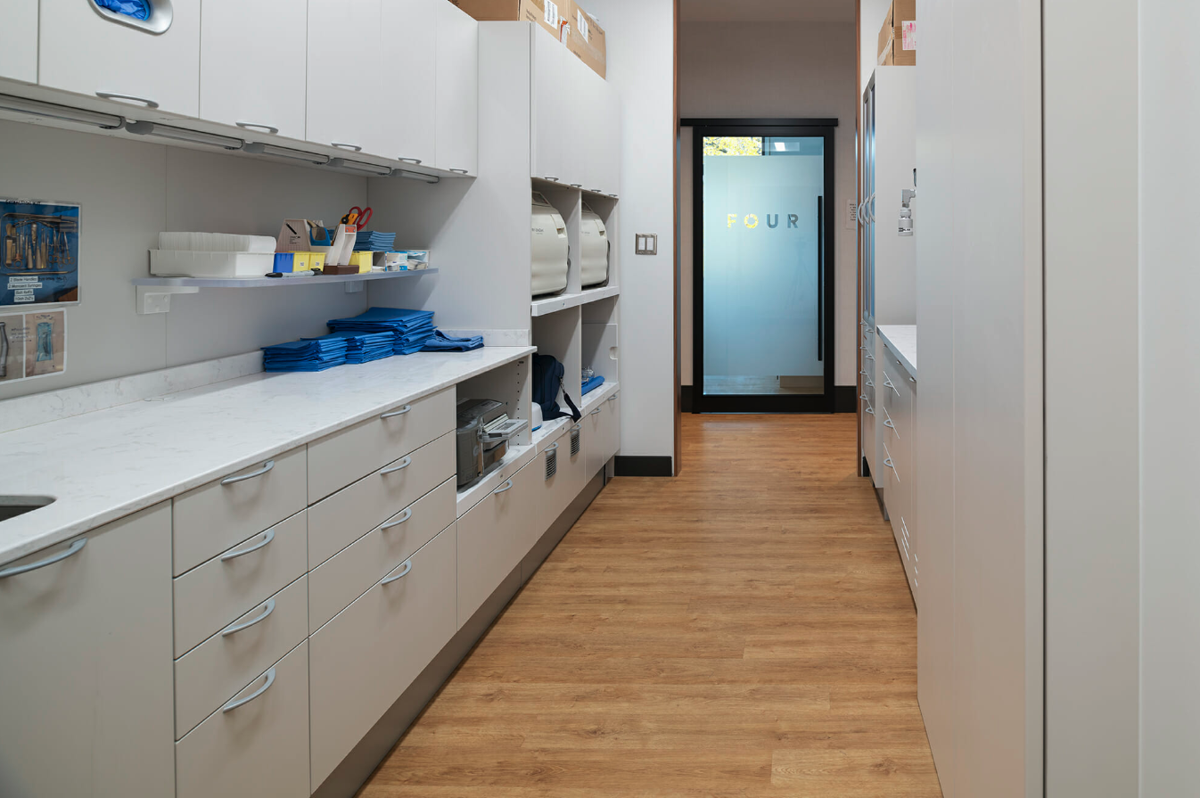 A wide hallway in a medical clinic with white drawers and cupboards, and light wood flooring. 