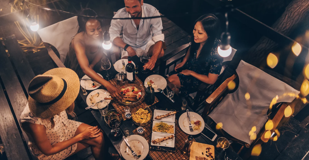 (A group of friends enjoying a cozy outdoor dinner under string lights, sharing drinks and food in a relaxed, intimate setting.)