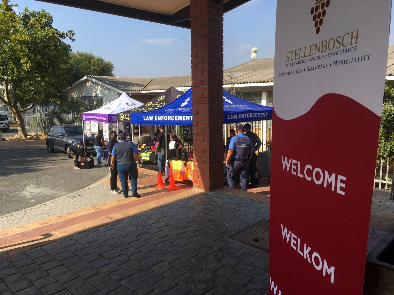 Members of the South African Police Service (SAPS), StellCARE, and neighbourhood watch stand under gazebos outside Eikestad Hall in Cloetesville.