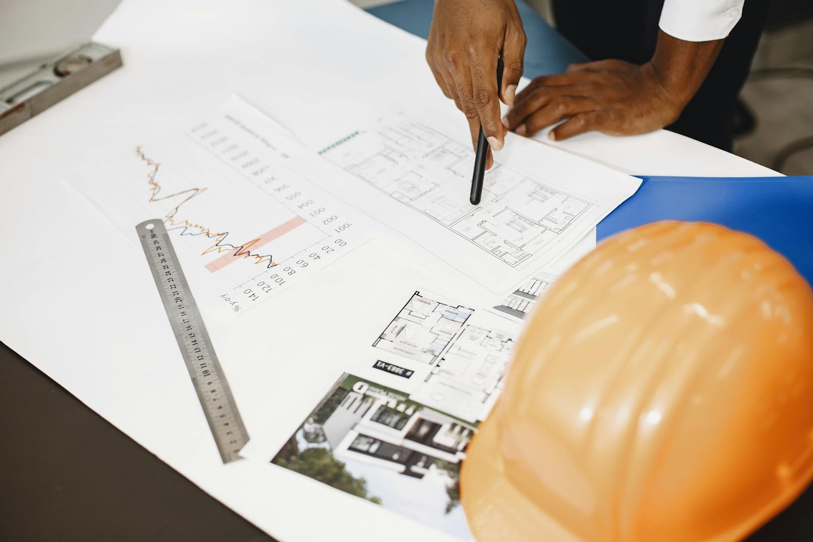A person points at architectural plans on a desk, surrounded by tools of their trade—a ruler, graph, orange safety helmet, and a blue folder. Architects find inspiration in the scattered building designs and photographs as they navigate their career path using BIM 3D Design.