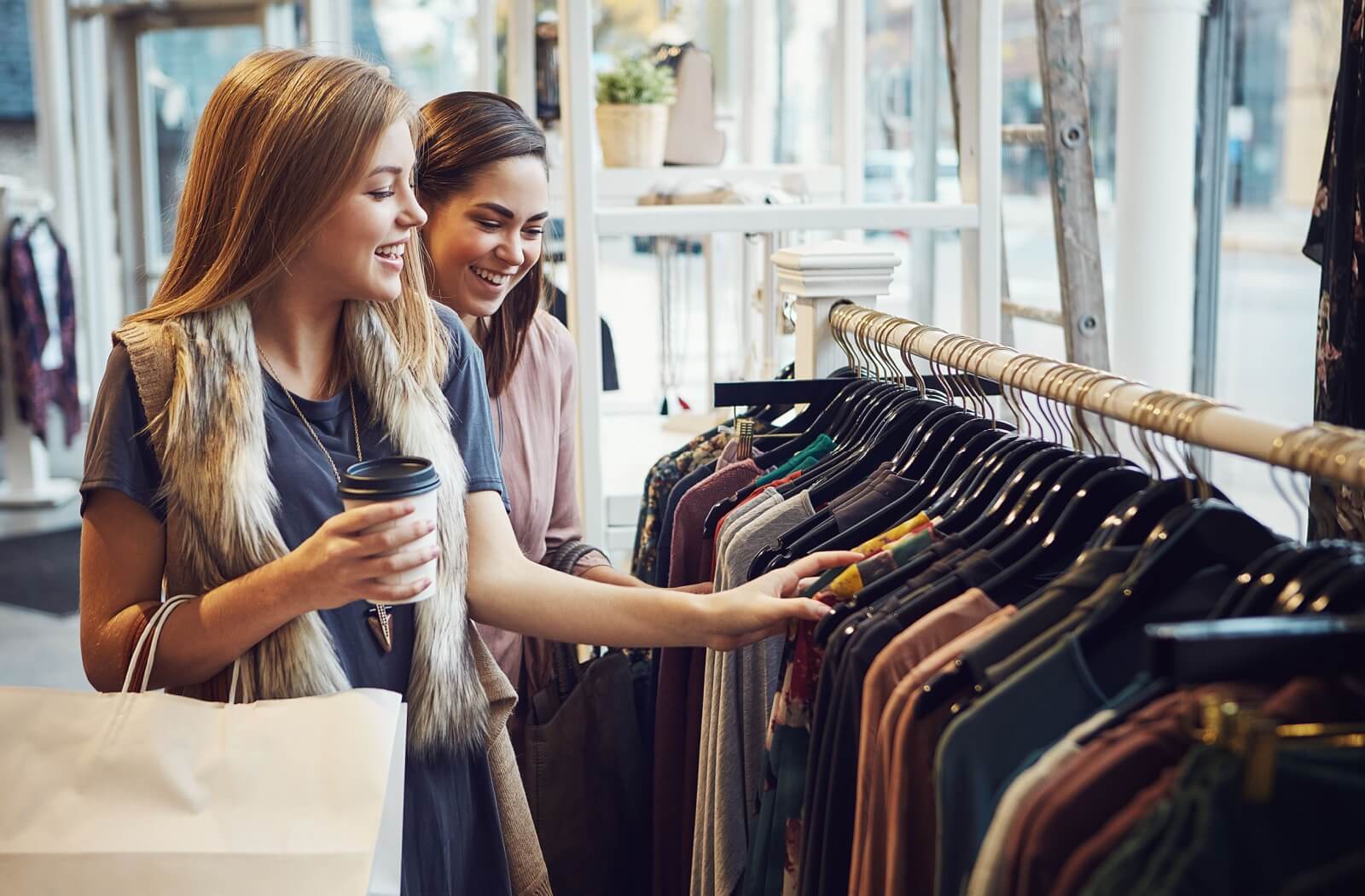 Two people holding coffees browse through racks of fashionable, second-hand clothing in a local consignment store.