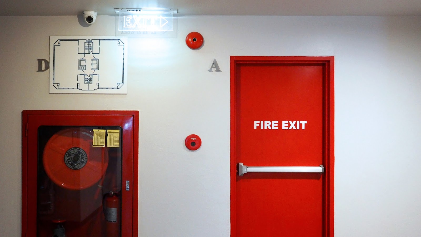 Red emergency exit door with push bar next to a fire hose cabinet and building evacuation map.