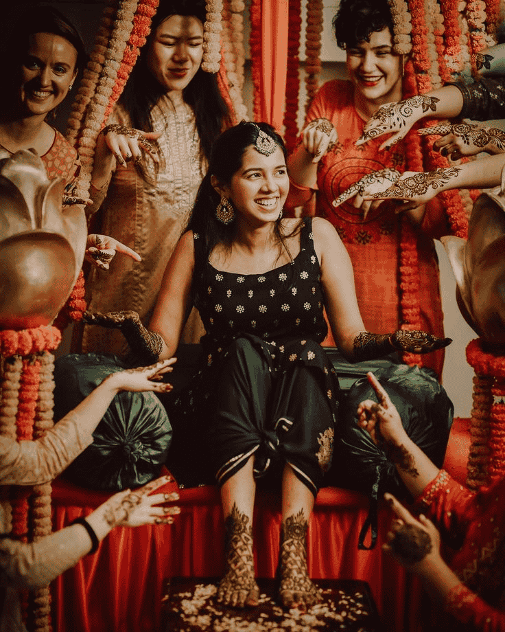 Mehndi photoshoot of a bride sitting while her mehendi is applied, with friends around her.