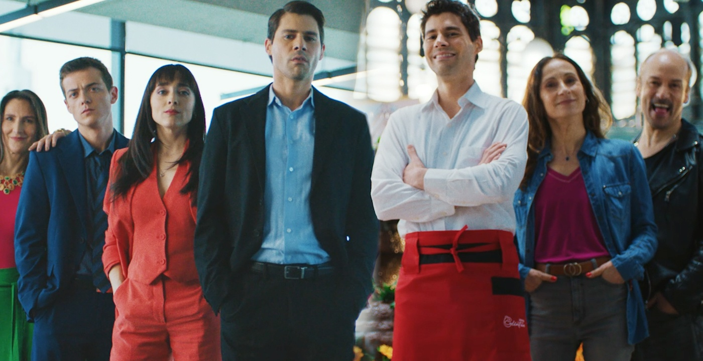 Grupo de personas posando en un ambiente moderno, donde un hombre viste un delantal rojo junto a otros personajes con diferentes estilos de vestimenta formal y casual.