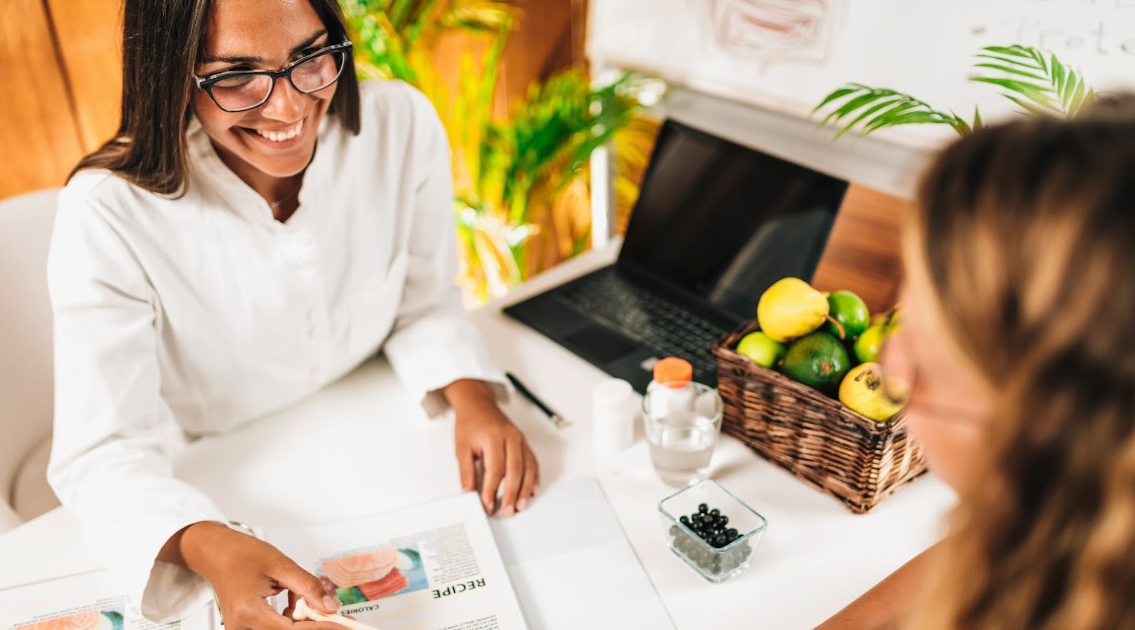 A smiling nutritionist-dietitian, wearing a white coat and glasses, is discussing with a client. They are seated at a table where the nutritionist is showing recipes in a binder. On the table, there is a basket of fresh fruit, a glass of water, dietary supplements, and a laptop. The environment is bright and welcoming, suggesting a consultation focused on balanced and healthy eating.