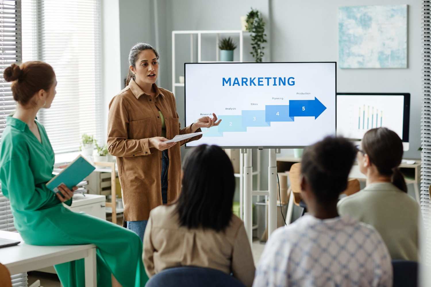 A woman presenting a marketing strategy to a group of colleagues in a modern office setting. She stands in front of a screen displaying a step-by-step marketing process, including research, analysis, ideas, planning, and production, while her team listens attentively