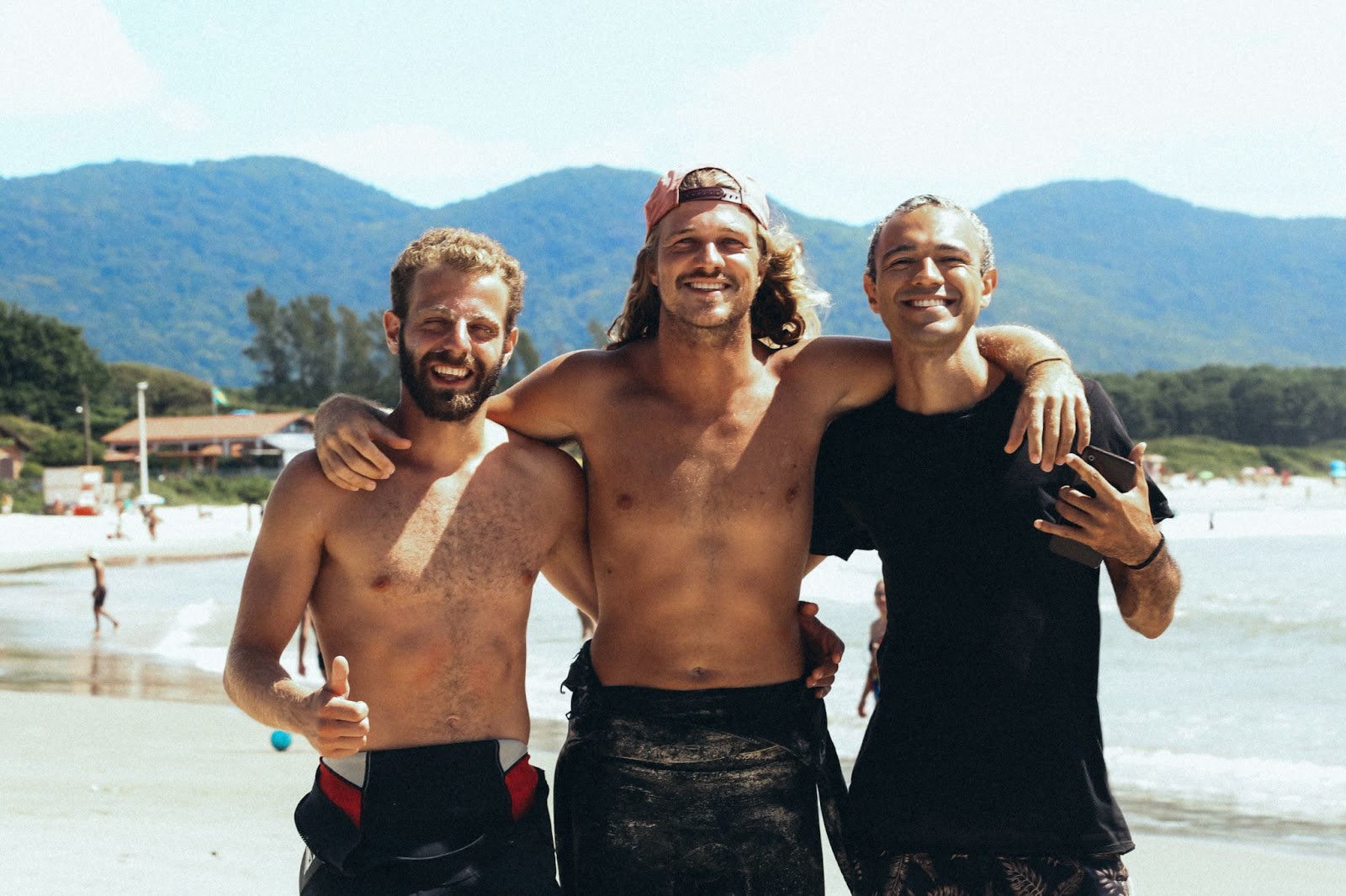 Três amigos abraçados sorrindo para a foto, na praia de Florianópolis 