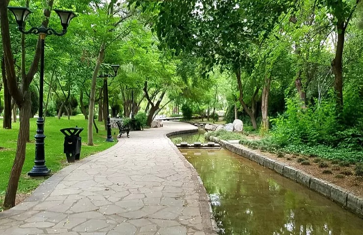 Peaceful view of Mellat Park in Tehran, Iran, featuring a stone pathway lined with trees, green grass, park benches, and a calm waterway, offering a serene urban escape.