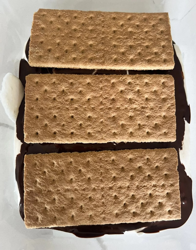 These frozen protein s'mores bars are gluten free, dairy free, high protein, can be low sugar, the perfect summer dessert. On a cutting board are the s'mores bars before cutting. Three golden brown graham crackers with hardened rich chocolate showing through the cracks between on the sides. A creamy, thick white protein whip shows spilling over the sides. Like ice cream sandwiches and ready to eat