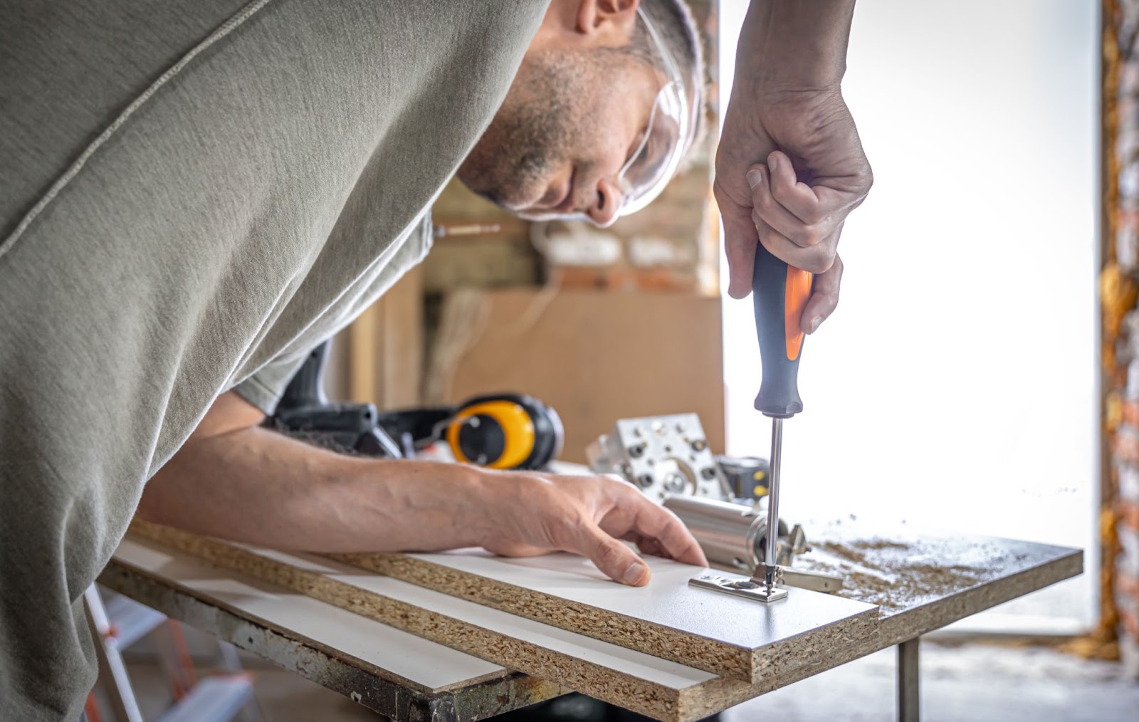 A handyman in a gray shirt, wearing protective glasses, carefully screws a self-tapping screw into wood.