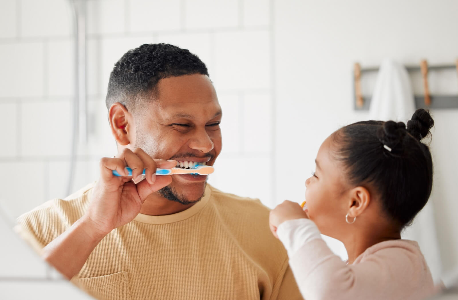 A parent smiling at their young child while brushing their teeth together and teaching them to avoid brushing too much.
