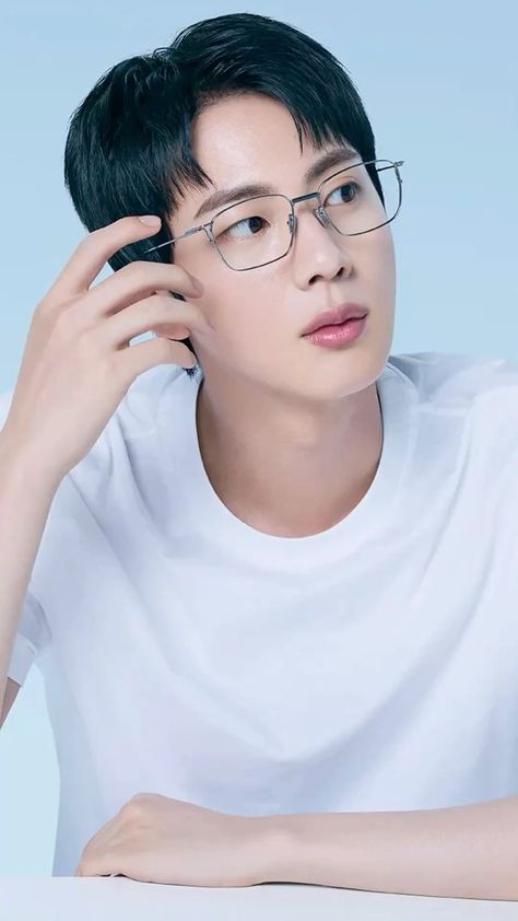 This contains an image of BTS Jin wearing glasses sitting at a table with his hand on his head and looking off to the side