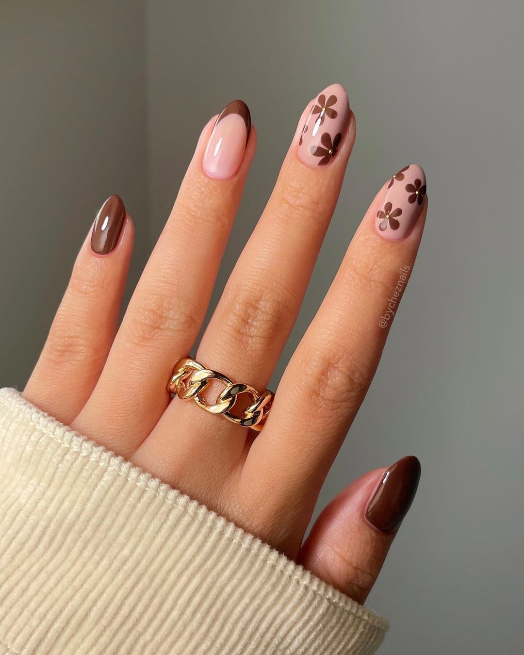 Two hands with long, almond-shaped, taupe-colored nails resting on a fluffy white surface.

