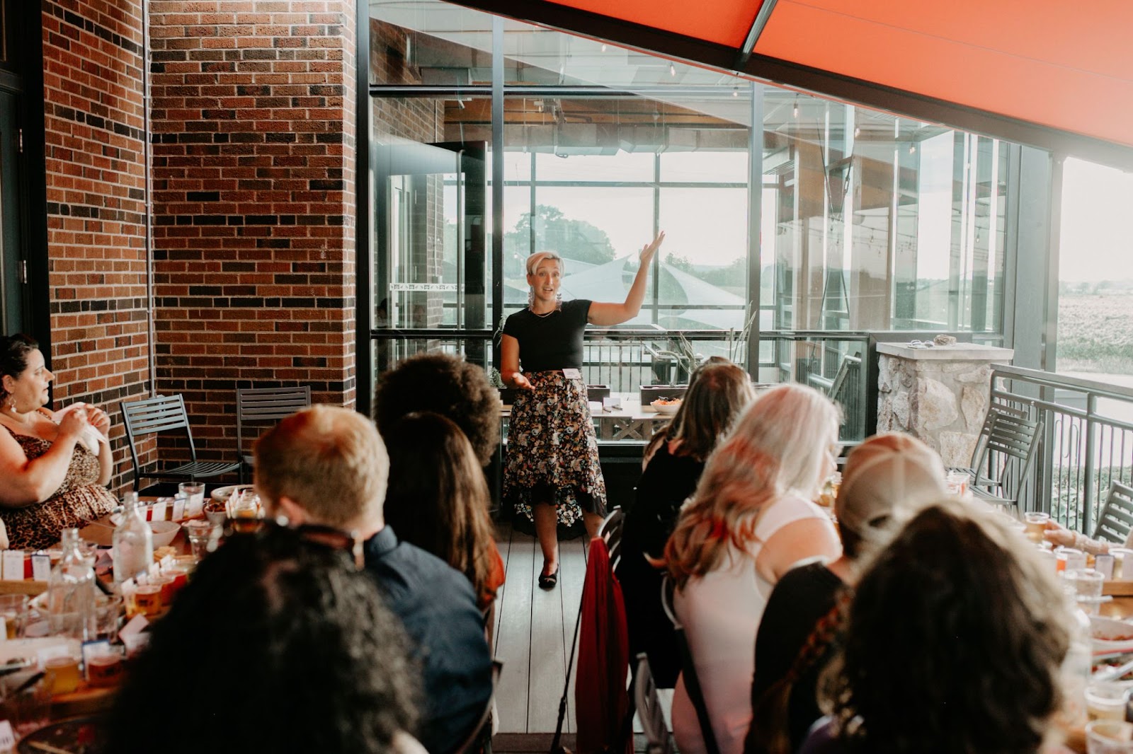 Tarzan is standing and giving a presentation to a seated audience in a room with brick walls and large windows. The presenter, wearing a black top and a floral skirt, has one hand raised and is speaking animatedly. The audience members, some of whom are visible from the back, are attentively listening.