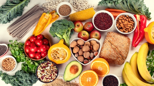 A colourful assortment of fruits, vegetables, nuts, and grains, including cherry tomatoes, broccoli, apples, bananas, oranges, avocados, almonds, whole-grain bread, corn, bell peppers, and kale leaves, arranged on a white table.