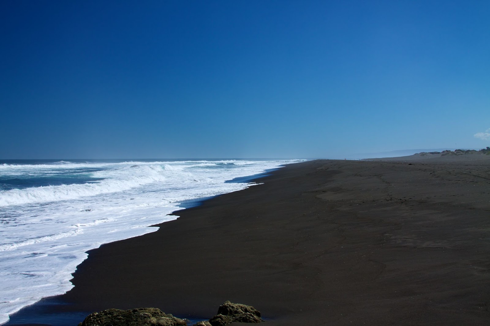 Dark sand beach with blue water and a clear blue sky overhead.