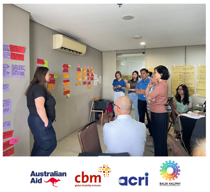 A photo of Kirsten Bate, Senior Programme Coordinator from CBM Australia, in front of a wall featuring metacards, and facing the Rise and Thrive program team during a discussion.