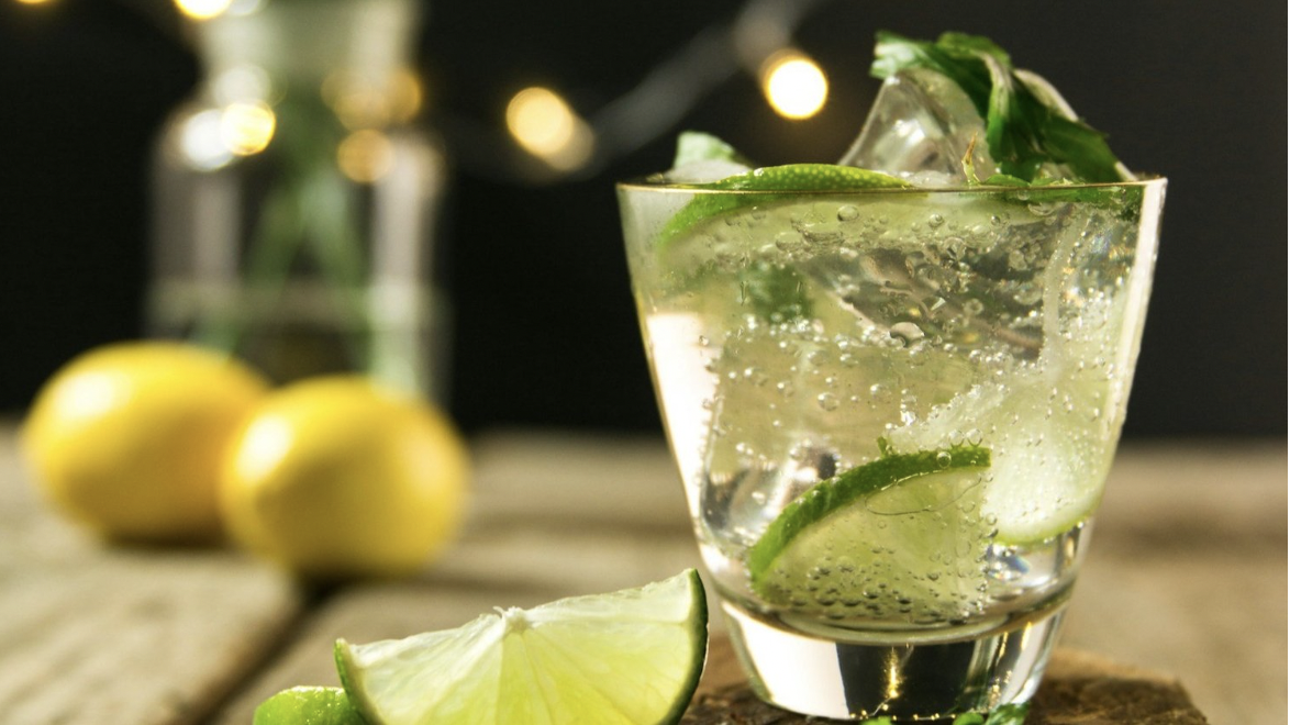 A glass is full of ice, mint, lime, rum, and soda water, and there are lemons in the background