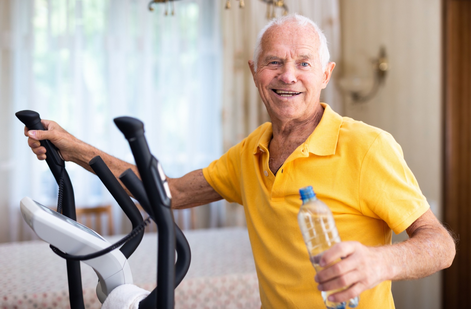 Senior man using an elliptical machine while holding a water bottle, exercising in a small indoor space.