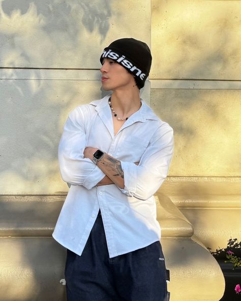 This contain Beomhan  with his arms crossed standing in front of a stone wall wearing a white shirt and black beanie