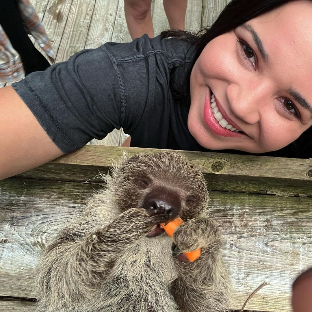 A woman smiling as she takes a selfie with a sloth eating a carrot.