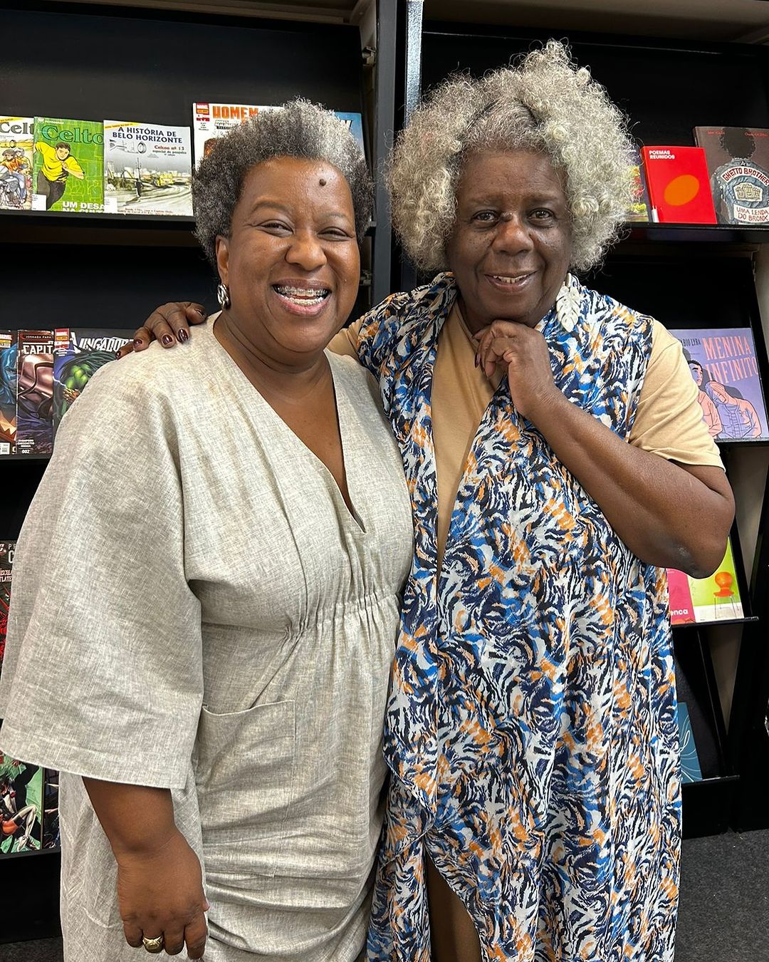 Macaé Evaristo, mulher negra de cabelo crespo, curto e grisalho, está usando aparelho e tem um sorrio largo. Ela está usando um macacão cinza. Ao seu lado, Conceição Evaristo, também com o cabeo crespo e grisalho, de tamanho médio, sorri para a foto. Ela usa uma roupa colorida