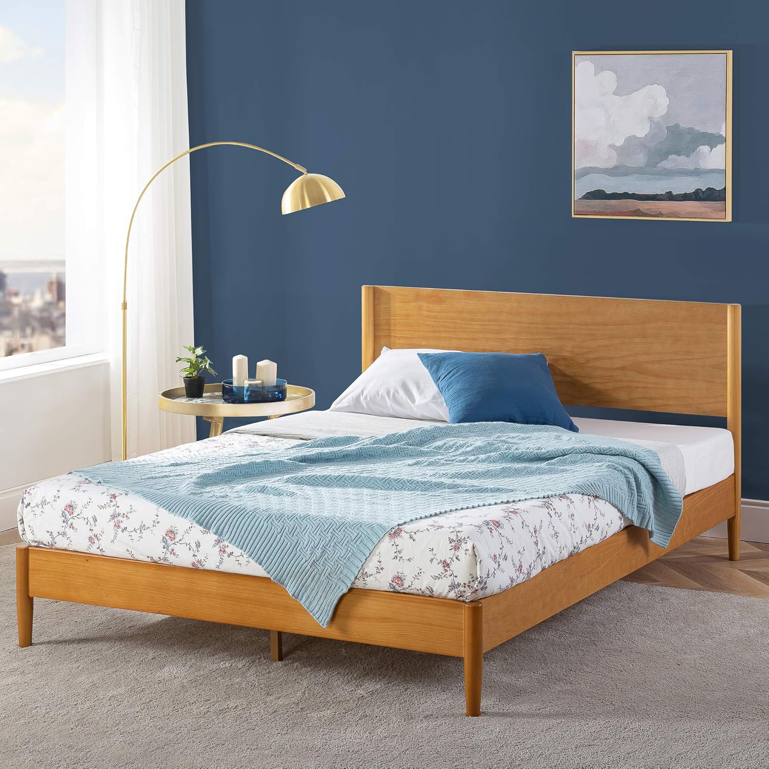 A minimalist wooden bed frame with a light blue blanket and pillows, paired with a modern arc floor lamp beside it.