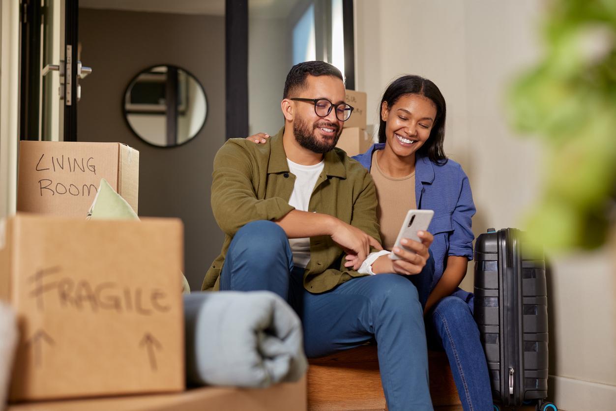 A young couple looking at their phone in front of their new home surrounded by boxes. 