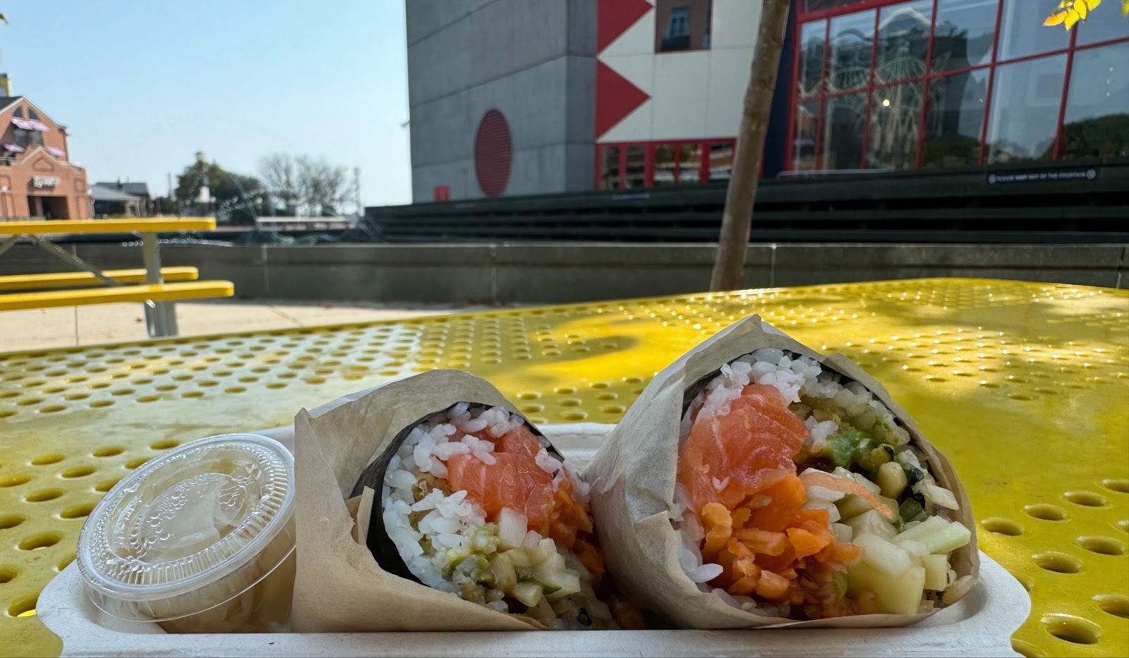 Sushi burrito in a container on top of a yellow table outside.  