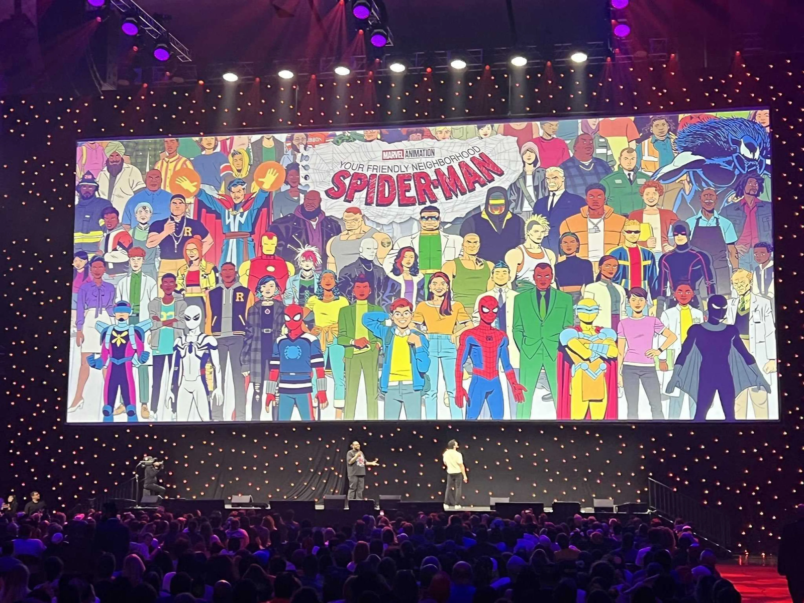 Presentación en un evento con una pantalla gigante que muestra una ilustración grupal de personajes del universo Spider-Man, destacando a varios héroes y villanos, mientras una multitud observa desde el auditorio.