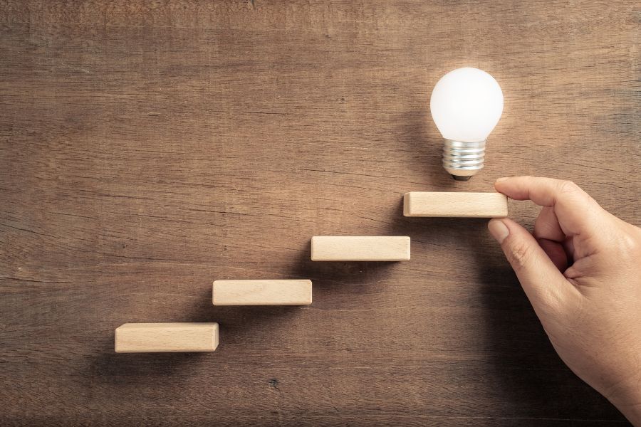 Hand arranging wooden steps leading to a glowing light bulb, symbolizing ideas.