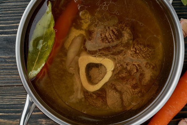 A pot of rich bone broth simmering with a beef marrow bone, bay leaf, and vegetables