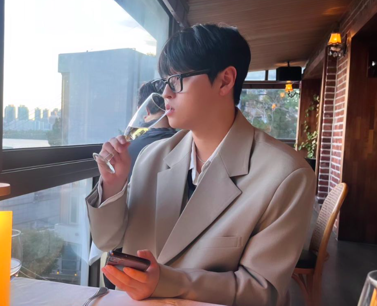 This contains an image of Gi Taek  wearing a brown suit,holding a glass cup and drinking wine