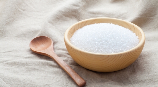 A wooden bowl filled with granulated white sugar, placed on a beige cloth, with a wooden spoon beside it.