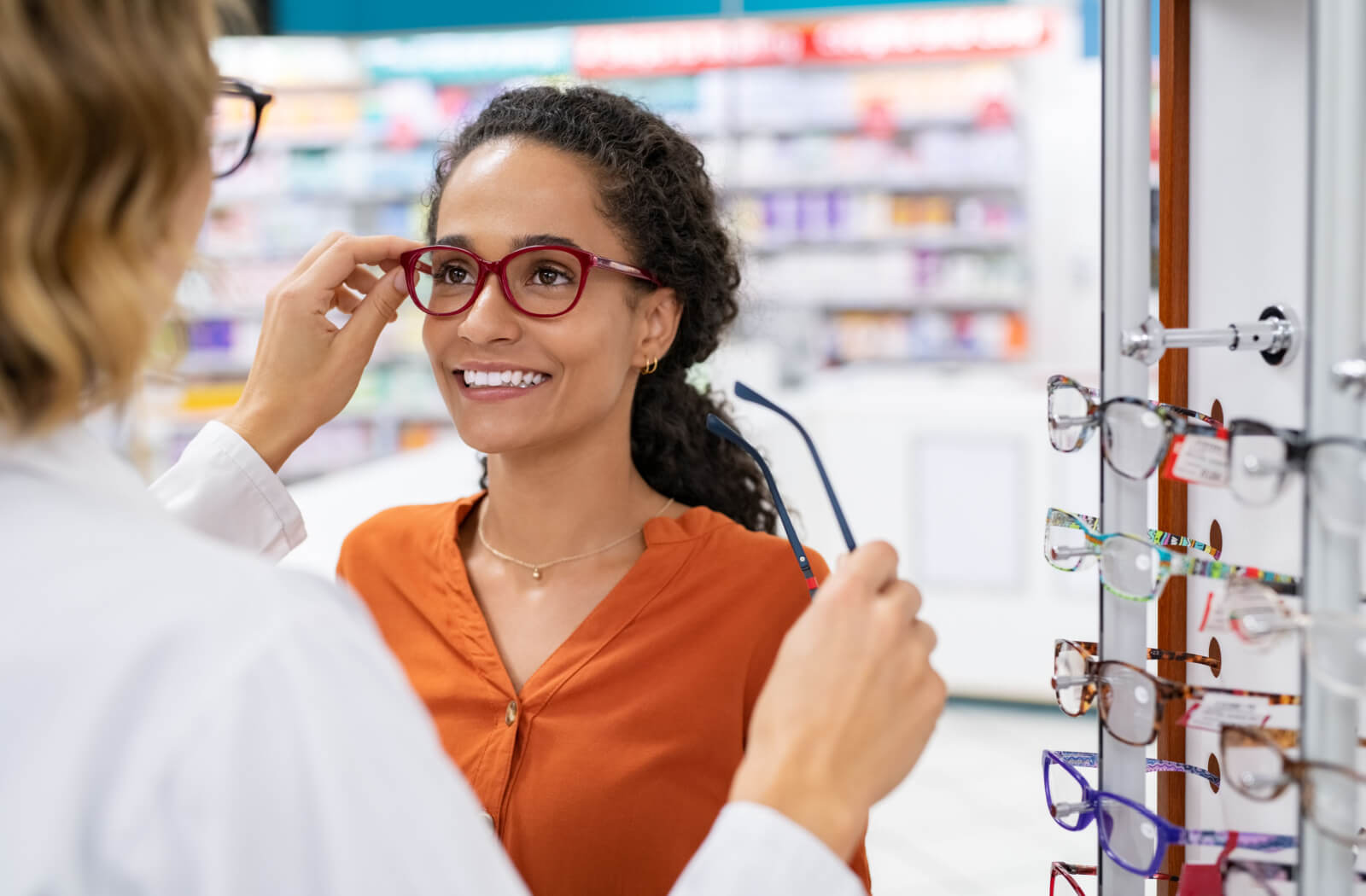 An optometrist helping a smiling patient choose a new pair of stylish glasses from a display wall.