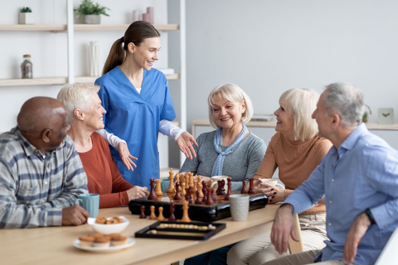 a group of seniors sit around a table with a chess board and a carer speaks to them