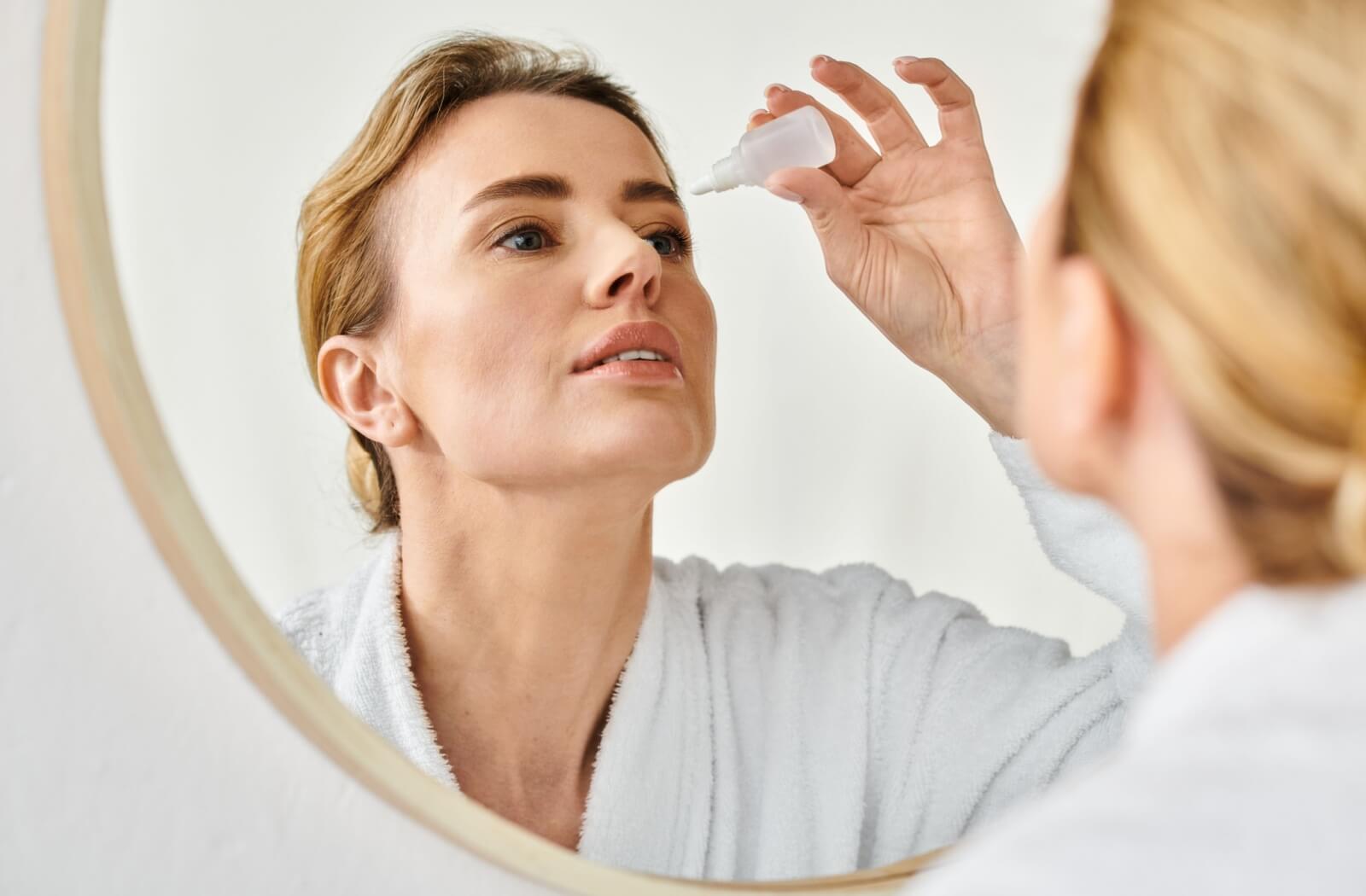 An adult in a white bathrobe applies eyedrops to their eyes in preparation for using an eye mask to relieve dry eye symptoms