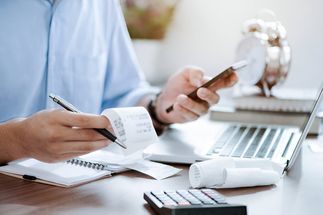 Person managing receipts and financial documents while using a smartphone and laptop for bookkeeping and tax preparation.