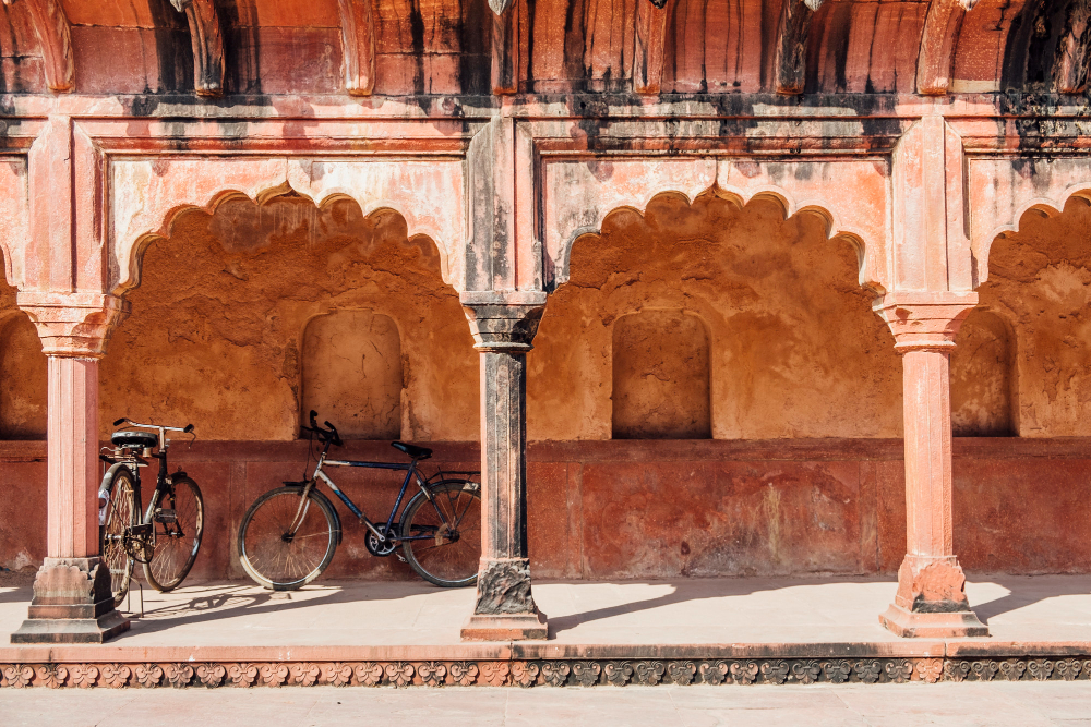 Two bicycles rest under the arches of an ancient terracotta-colored building. The structure, echoing vernacular wisdom, features ornate, curved arches and weathered walls, with sunlight casting shadows on the stone floor.