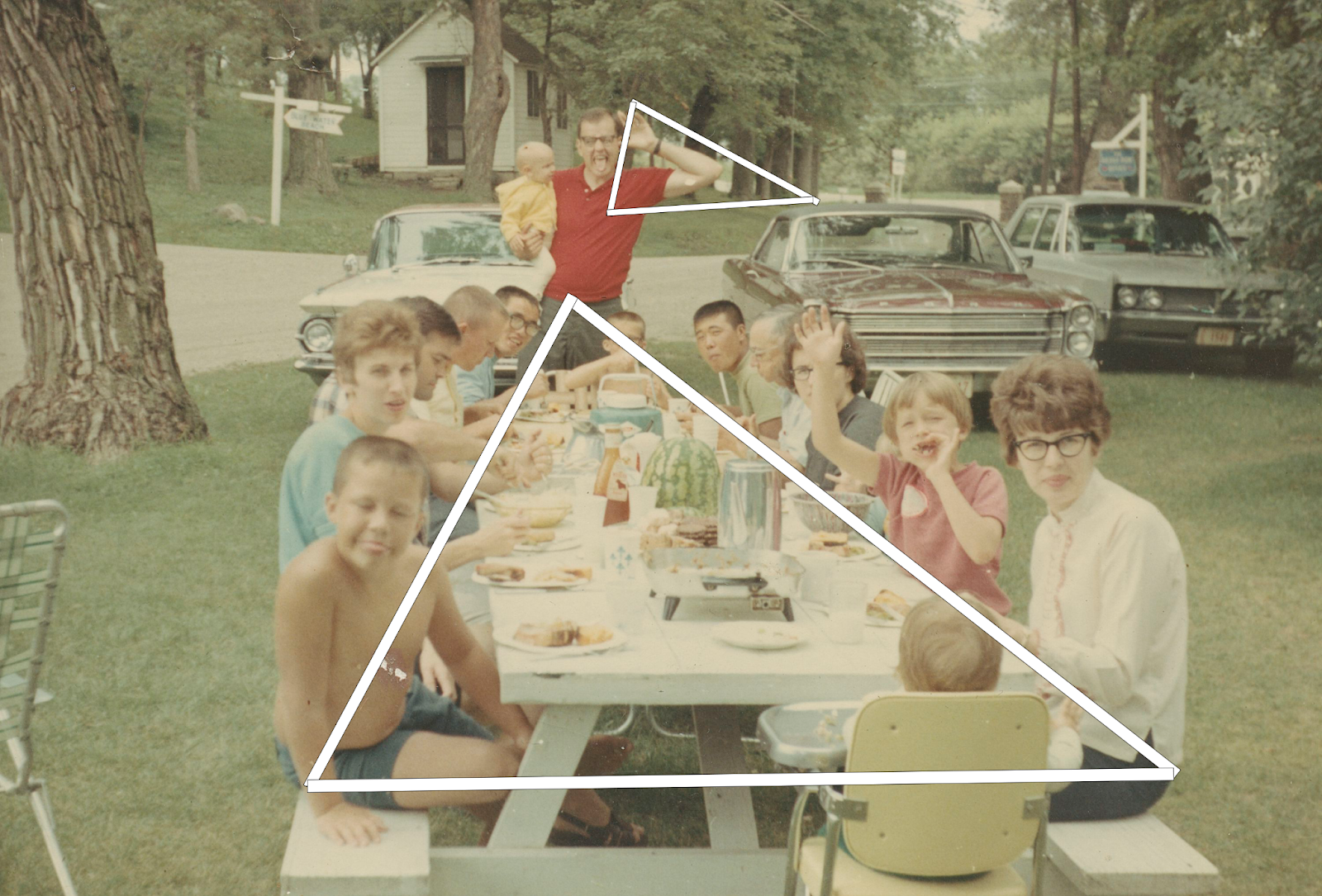 A color photo depicting a long picnic table with many individuals, adults & children. eating a meal. A white triangle connects the sides of the table and another connects a man's outstretched elbow to a car nearby