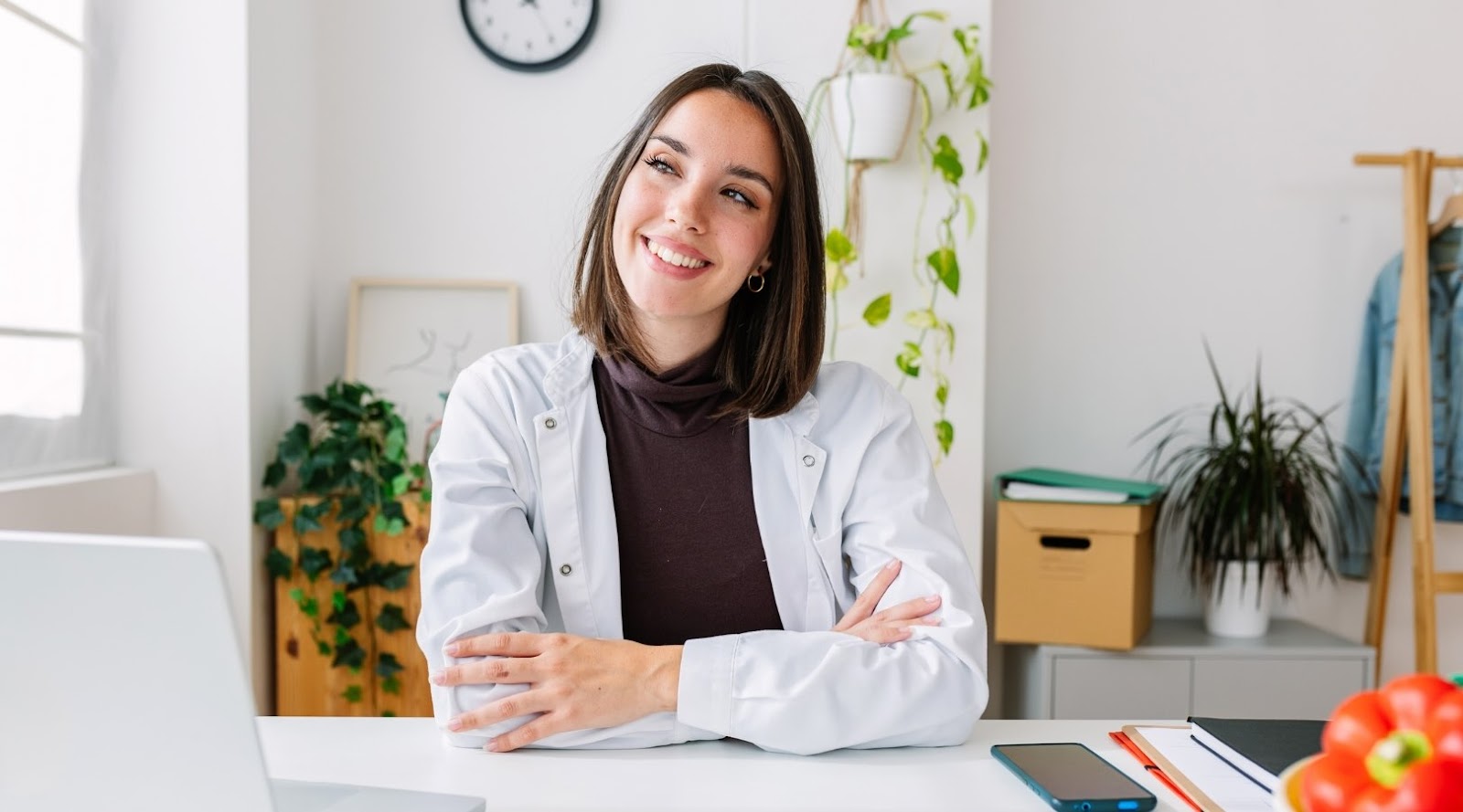 A smiling young nutritionist-dietitian, wearing a white coat, is seated at her desk with her arms crossed, looking thoughtful and positive. In front of her, a laptop is open, and on the table, there's a phone, a notebook, and a bowl of fresh fruit, mainly strawberries and tomatoes. The environment is bright and green, with plants in the background, suggesting a healthy and inspiring workspace.