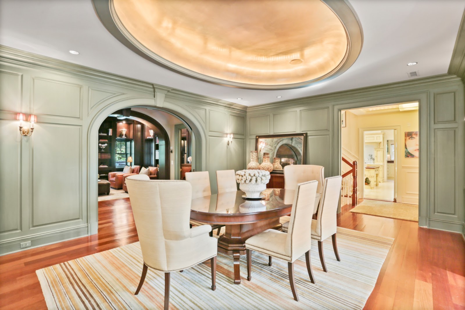 A custom painted paneled dining room in soft sage green features an oblong mahogany table, upholstered chairs and oval recessed ceiling.