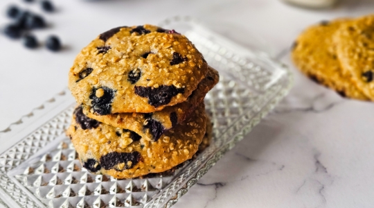 Une pile de biscuits aux bleuets, amandes et chocolat noir sur une assiette en verre, avec un verre de lait et des bleuets frais en arrière-plan.